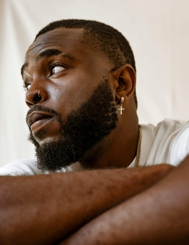 Gros plan d’un homme afro, barbe afro soignée et texturée, fond clair, boucle d’oreille en croix.