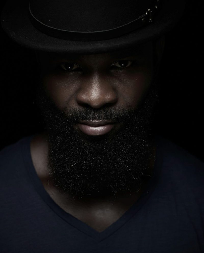 Portrait d’un homme noir à la barbe dense, chapeau noir, fond sombre, éclairage studio.